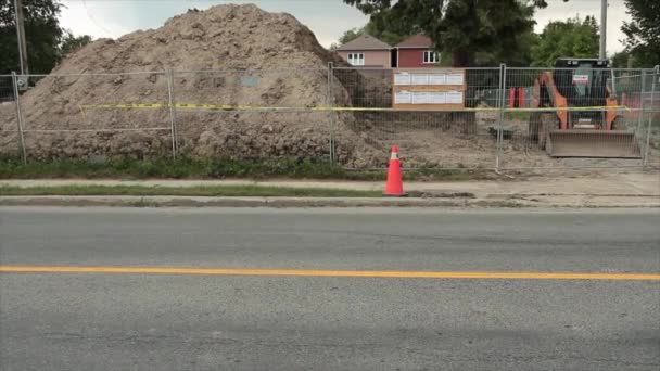在银色栅栏后面堆积如山的泥土 墙上贴有建筑许可证 旁边停着黄色的黑色建筑车辆 前面的道路经过 后面是住宅 — 图库视频影像