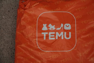 Temu turuncu dikdörtgen çanta paketi üzerinde logo var. Kaldırım çimento üzerinde duruyor. Sol çerçeve, logo merkezi.