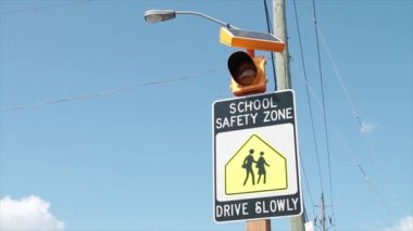 Okulun güvenli bölgesi yavaş yavaş imzalayın. Üzerinde mavi gökyüzü, siyah beyaz ve sarı renkli insanların yürüdüğü ve yanıp sönen sarı ışık var.