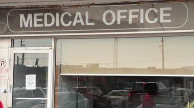 Tıp bürosu, tıp bürosunun vitrininde pencere ve kapı işaretleri yazıyor, genç, orta yaşlı, siyah bir erkek geçiyor.