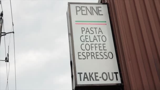 イタリアンレストランは 店頭から突き出る白い背景のテキストに黒い筆記テキストでペンネパスタジェラートコーヒーエスプレッソを言う垂直長方形のサインを取り出します — ストック動画