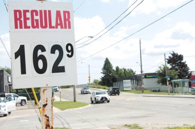 Beyaz dikdörtgen arkaplanda siyah ve kırmızı yazılı normal benzin fiyatı 162 9. Arka planda yol ve köprü ve araç arabaları ile birlikte.