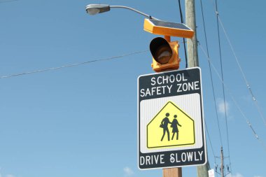 Okulun güvenli bölgesi yavaş yavaş imzalayın. Üzerinde mavi gökyüzü, siyah beyaz ve sarı renkli insanların yürüdüğü ve yanıp sönen sarı ışık var.