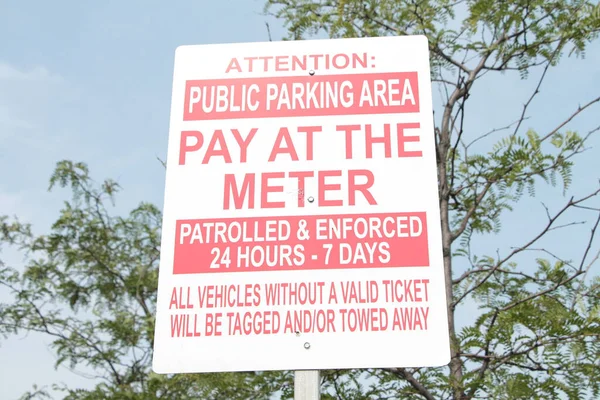 attention public parking area pay at the meter patrolled and enforced 24 hours 7 days all vehicles without valid ticket will be tagged and or towed rectangle sign with tree and sky behind
