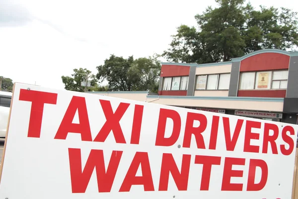 taxi drivers wanted sign in red capital letters writing text with buildings and trees behind