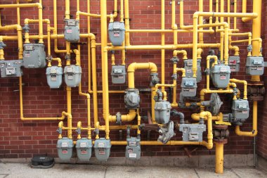Bir sürü gri gaz sayacı bir araya toplanmış tuğla duvara monte edilmiş ve içinden sarı gaz boruları çıkıyor.