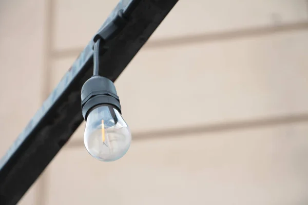 small exterior outdoor string light bulb with black base hanging upside down mounted to metal bar post turned off with wall behind
