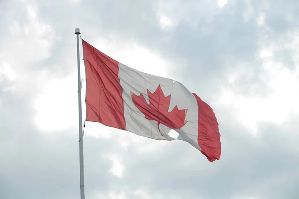 large canadian canada flag on flagpole with rip hole in it blowing waving in wind with sky clouds sun behind shining