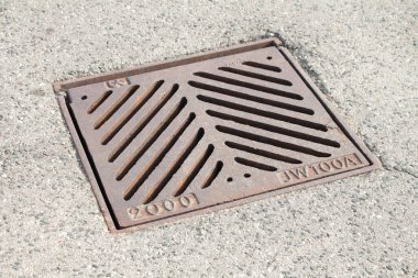 Üzerinde çapraz çapraz çukur çizgileri olan kuru asfalt arasında kahverengi kare kanalizasyon. Yazın dış kısımda 2000 jw100a yazıyor. Üzerinde güneş var.