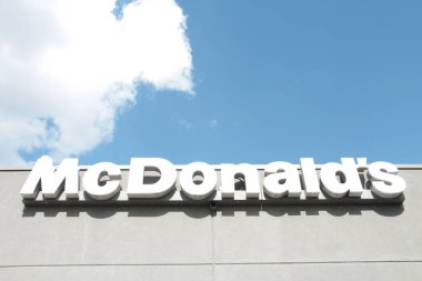 McDonalds, gri restoran duvarına beyaz harflerle işaret logosu yazıyor. Arkasında bulutlar var. Yazın üzerinde güneş parlıyor.
