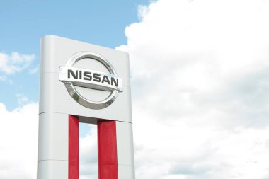 Nissan gümüşü logo açık havada beyaz kırmızı dikdörtgen sütun arasında boşluk ve bulutların arkasında gökyüzü.