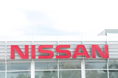 Nissan kırmızı logosu dışarıdaki bayiliğin önünde, pencerenin altında, arkasında gökyüzü olan bir tabela.