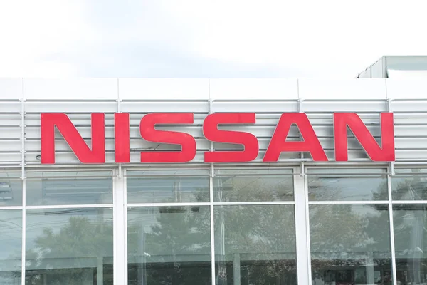 Nissan kırmızı logosu dışarıdaki bayiliğin önünde, pencerenin üstünde, arkasında gökyüzü olan orta çerçeve.