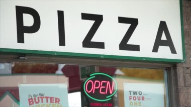 Pizza sözcüğü başlık yazma tabelası pencerenin üstünde. Yazın dışarıdaki mağaza vitrinindeki pizzacıda yanıp sönen neon açık işareti var.