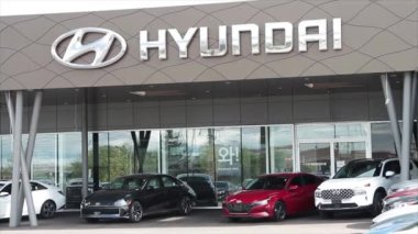 Gümüş logolu hyundai bayisi. Gri bayiliğin ön duvar cephesinde yazılar yazıyor. Pencerelerin ve park halindeki arabaların ve arabaların dış cephelerinin üzerinde yansıması var.