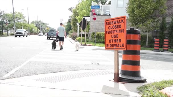 人行道上封闭的行人使用其他人行道 在人行道上写着标题文字橙色的黑色矩形建筑标志 人行道上有遛狗的人和在路上经过的车辆 — 图库视频影像