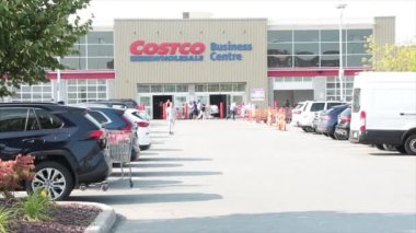 Costco toptan satış merkezi. Yaz mevsiminin ön tarafında park yeri ve ağır müşteri trafiği var. Bir kişi dükkandan uzaklaşırken diğeri de oradan geçen araba ve el arabalarıyla.