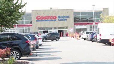 Costco toptan satış merkezi. Yaz mevsiminde otoparkın önünde bir araç kamyonu park yerinden geçiyor ve müşteriler yürümeye devam ediyor.