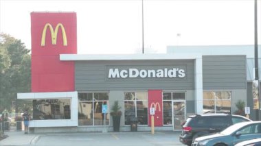 McDonalds restoranının girişi park yeri ve önünde arabaların önünde telefon ve araba önünde duran kişi McDonalds 'ın camlarında yansıması ile önünden geçiyor.