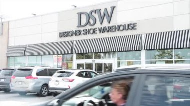 DSW tasarım ayakkabı deposu ön girişi. Arabalar park edilmiş ve geçiyor. Dışarıdaki mağazanın önünde siyah ile işaret logosu yazıyor.