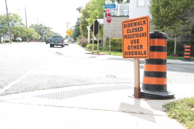Kaldırımlar kapalı yayalar diğer kaldırımları kullanır. Yazı başlığı: Turuncu siyah dikdörtgen inşaat tabelası kaldırım kenarındaki trafik delinatörü direğinin önündeki yol kenarına.