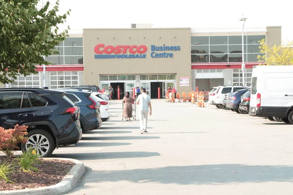 Costco toptan satış merkezi. Yazın park yeri önünde iki müşteri var. İnsanlar alışveriş arabasıyla dükkana doğru yürüyorlar.