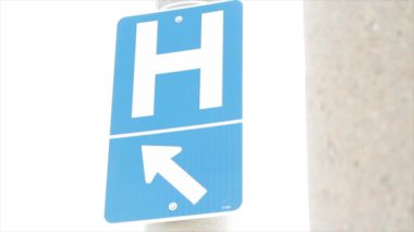 H harfi, büyük harf h harfi üzerinde mavi beyaz dikdörtgen işareti ve sol kuzeybatıyı işaret eden ok açık havada mavi dış tarafta, güneş arkadan vuruyor, açıyla vuruyor.