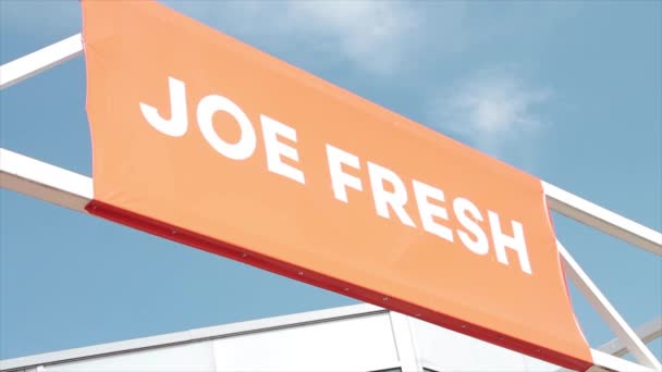 ジョー新鮮なブランドのロゴ広告の広告の旗の印 金属棒の横の長方形 オレンジの背景の白い書き込みの外側の空の雲の建物 — ストック動画