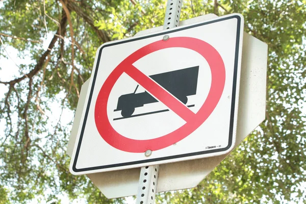 no trucks allowed square sign on metal post with stop sign behind and tree branches and sky