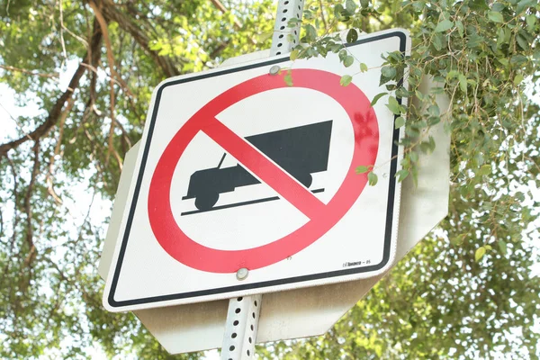no trucks allowed square sign on metal post with stop sign behind and tree branches leaves hanging in front and sky behind