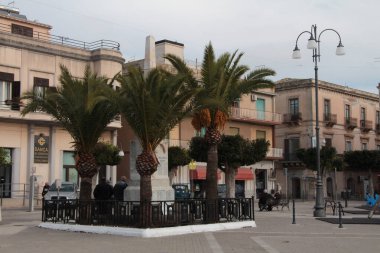 Pachino Sicilya 'daki İtalya Piazza Anıtı. Etrafında palmiye ağaçları ve arkasında da bina dükkanları var.