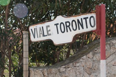 Pachino 'nun arkasında ağaçlar ve taş çitler olan Toronto Caddesi levhası. Sicilya usulü yazılar. Yazı: Beyaz, siyah, kırmızı.