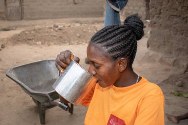Opialu, Benue State - 6 Mart 2021: Afro-Afrikalı Kadın Evinin dışındaki Paslanmaz Kupadan Turuncu Tişört İçen Su Giyiyor