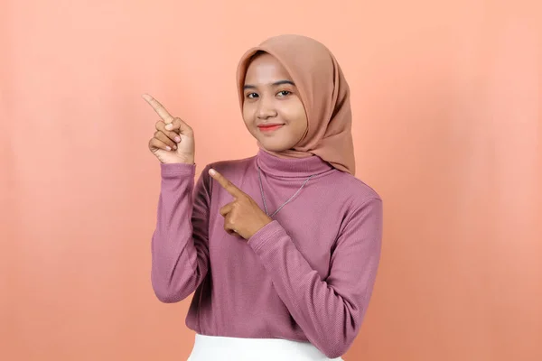 快乐的年轻的穆斯林亚洲女人指着紫色毛衣空旷的地方 与桃色背景隔离 — 图库照片