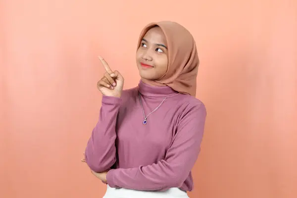 快乐的年轻的穆斯林亚洲女人指着紫色毛衣空旷的地方 与桃色背景隔离 — 图库照片