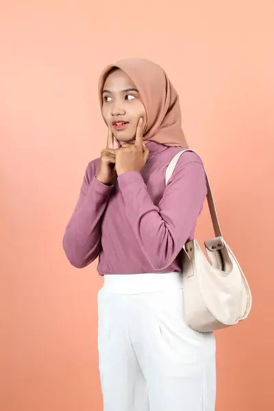 かわいい若いアジアのムスリム女性は 白いバッグを運んで紫色のセーターを着ている間 指で頬を握ります — ストック写真