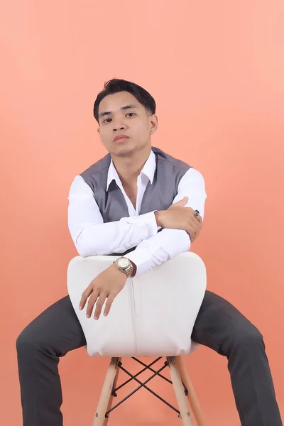 スーツのベストを着た若いアジアの実業家は自信を持って後ろから見た白い椅子の上に座っているカメラを見つめます — ストック写真