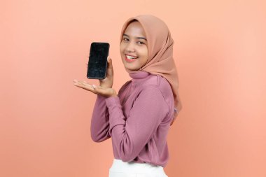 Güzel, genç Asyalı bir kadın elinde bir telefon tutuyor ve dişlerini gösteren bir gülümsemeyle telefonunu gösteriyor.