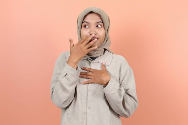 Güzel, genç Asyalı bir kadın bir eliyle ağzını kapatıyor ve diğer eliyle göğsüne şaşkın bir ifadeyle dokunuyor.