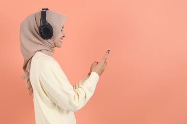 Genç Asyalı Müslüman kadın krem rengi abaya tesettür kulaklığı takıyor müzik dinliyor şeftali arka planda izole edilmiş cep telefonu kullanıyor insanlar İslami bakış açısı kullanıyor