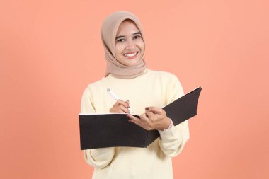 Krem rengi kazak giyen, elinde kalemle gülümseyen Asyalı güzel bir kadın şeftali ağacından izole edilmiş kameraya bakıyor.