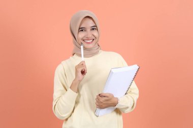 Kremalı süveter giyen neşeli Asyalı Müslüman kadın şeftali arkaplanından izole edilmiş, gülümseyen bir not defteri tutuyor.