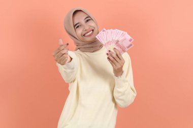 Krem rengi süveter içinde gülümseyen Asyalı Müslüman kadın elinde nakit para, 100 rupiah banknot, şeftali arka planda saklanmış el kol hareketi yapıyor. İnsanlar dini yaşam tarzı kavramları
