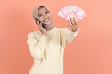 Heyecanlı, krem kazaklı, Endonezya rupia banknotlarından nakit para tutan ve cep telefonuyla el kol hareketi yapan Asyalı Müslüman bir kadın. İnsanlar dini yaşam tarzı konsepti