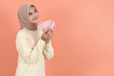 Krem rengi kazaklı güzel Asyalı Müslüman kadın elinde Endonezya rupia banknotlarıyla nakit para tutuyor. 