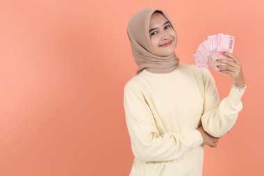 Endonezya rupia banknotlarından nakit para tutan ve para gösteren krem kazaklı güzel Asyalı Müslüman kadın. İnsanlar dini yaşam tarzı kavramları