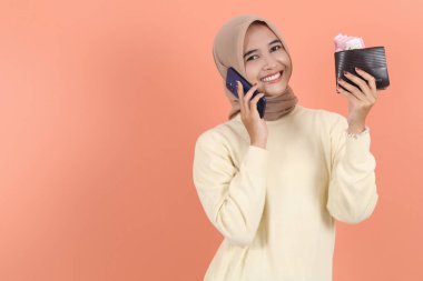 Krem rengi kazaklı Asyalı genç Müslüman kadın elinde cep telefonu ve elinde para dolu banknotlarla arıyor. Halk yaşam tarzı konsepti