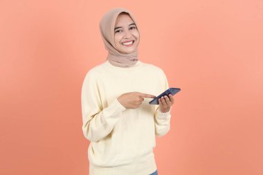 Krem süveter ve tesettürlü gülümseyen Asyalı Müslüman kadının portresi cep telefonu kullanıyor, ekrandaki boş ekrana işaret ediyor.