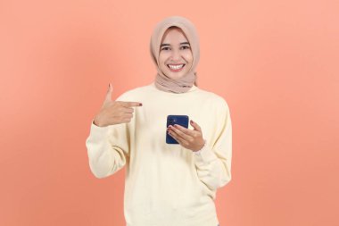 Krem süveter ve tesettürlü gülümseyen Asyalı Müslüman kadının portresi cep telefonu kullanıyor, ekrandaki boş ekrana işaret ediyor.
