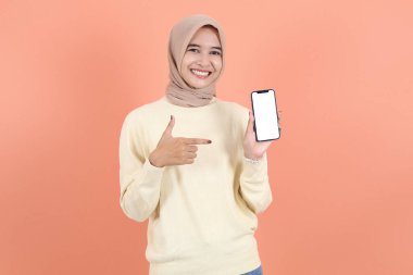 Krem süveter ve tesettürlü gülümseyen Asyalı Müslüman kadının portresi cep telefonu kullanıyor, ekrandaki boş ekrana işaret ediyor.